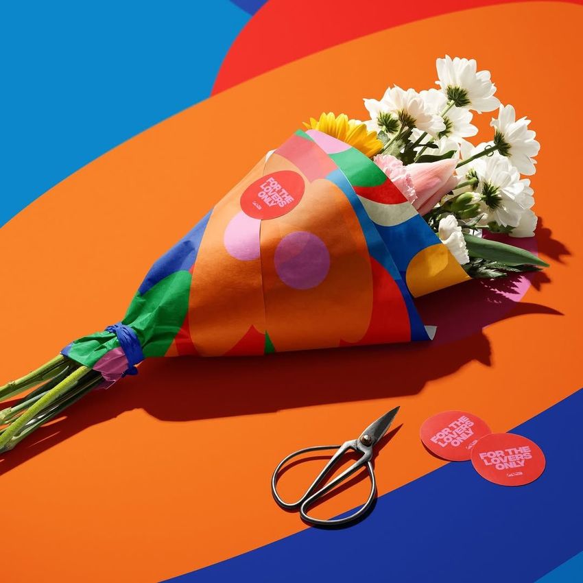 Colorful flower packaging mockup with white daisies and a pink rose wrapped in vibrant patterned paper on a bold abstract background.