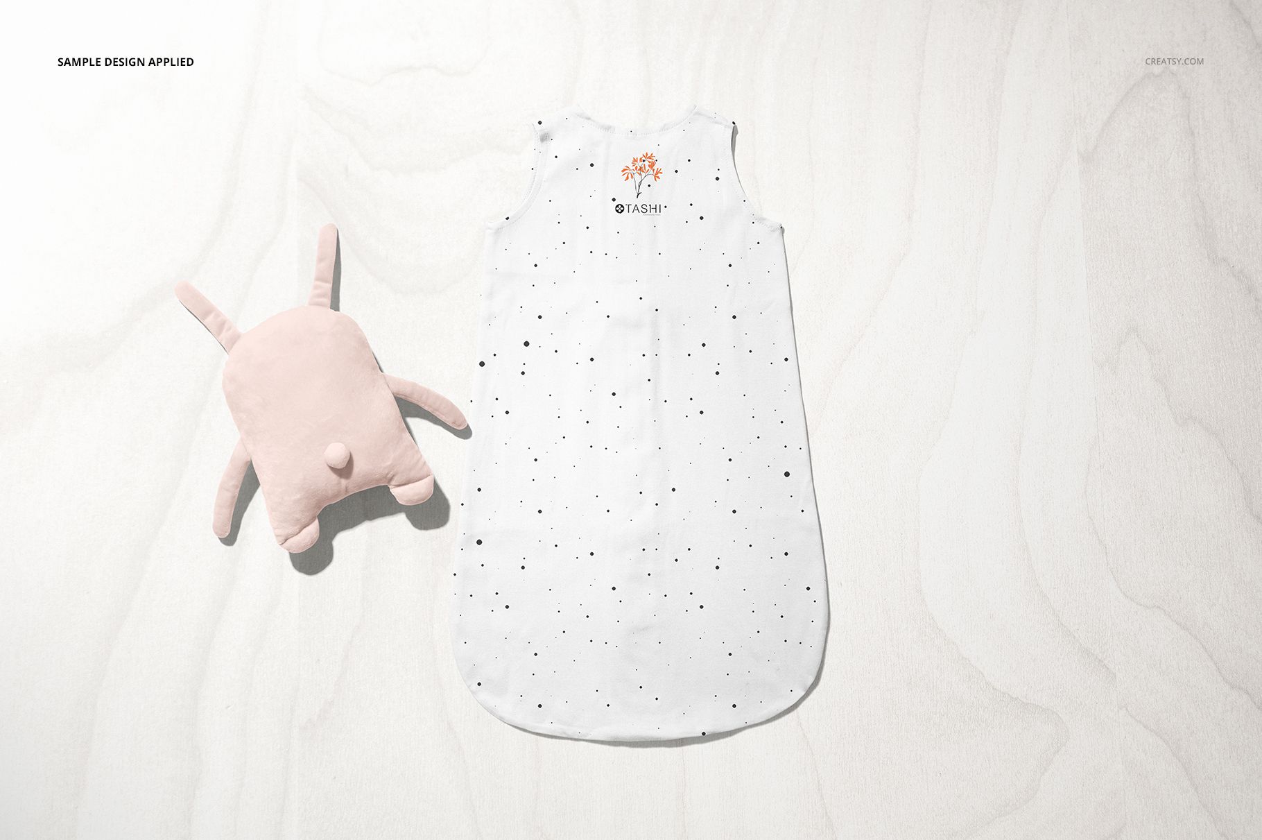 Top view of a baby wearable blanket mockup in white with small black speckles, accompanied by a light pink plush toy.
