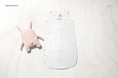 Top view of a baby wearable blanket mockup in white with small black speckles, accompanied by a light pink plush toy.