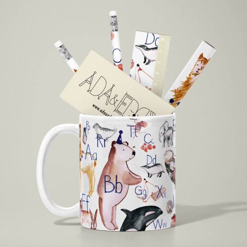 Mug mockup with illustrated animals and letters, filled with pencils, a ruler, and a card on a neutral background.