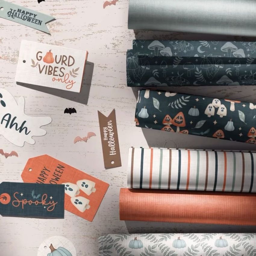 Halloween wrapping paper mockup showing several rolled sheets with ghost, pumpkin, and striped patterns on a light wood surface.