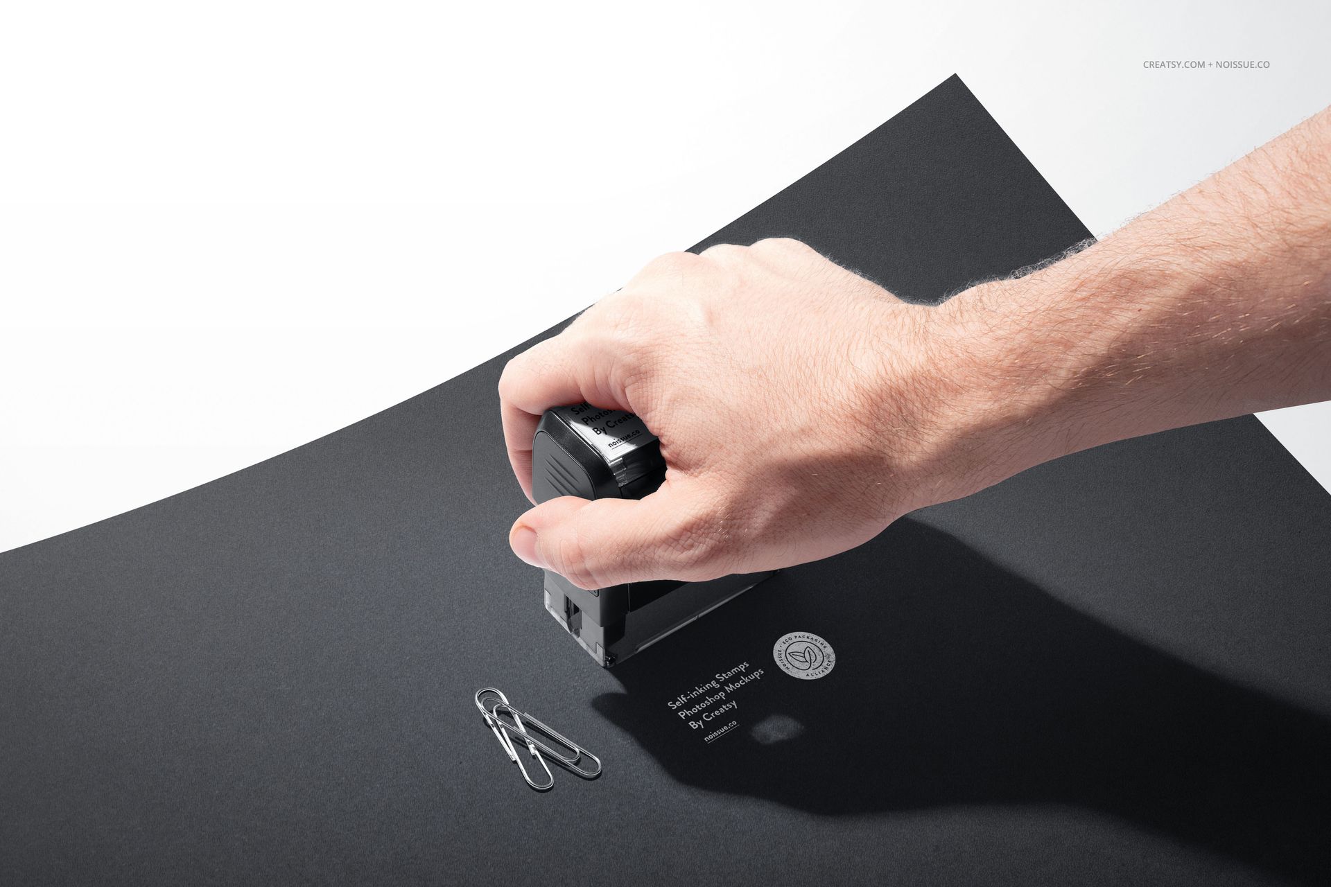 Close-up of a self-inking stamp mockup on black stationery, hand applying stamp near two metal paperclips.