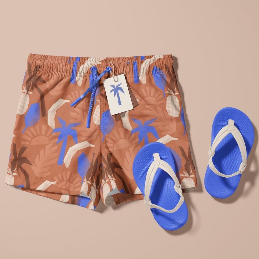 Kids swim shorts mockup with a tropical leaf and palm pattern in brown, blue, and beige, shown with blue sandals on a beige background.