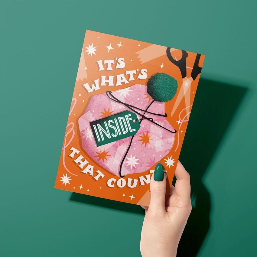 Greeting card mockup featuring a woman's hand holding a card with bold text, playful graphics, and an orange background.