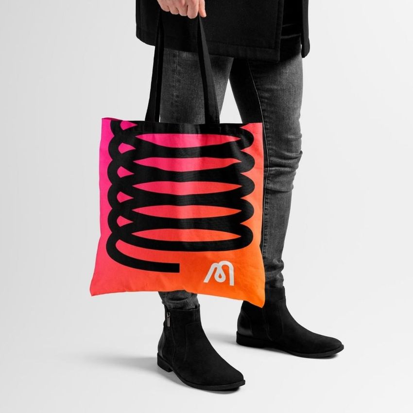 Tote bag mockup featuring a person holding a bag with a bold black abstract design on a pink and orange gradient background.