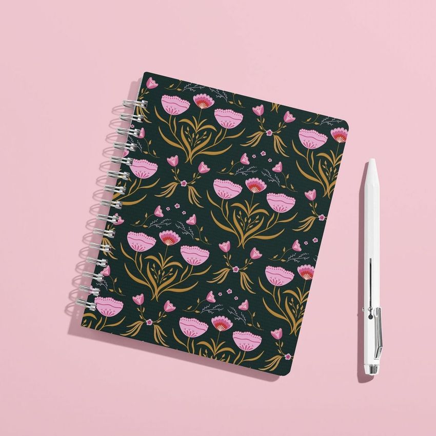 Coiled notebook mockup with a dark cover featuring a pink floral pattern, placed next to a white pen on a pastel pink surface.