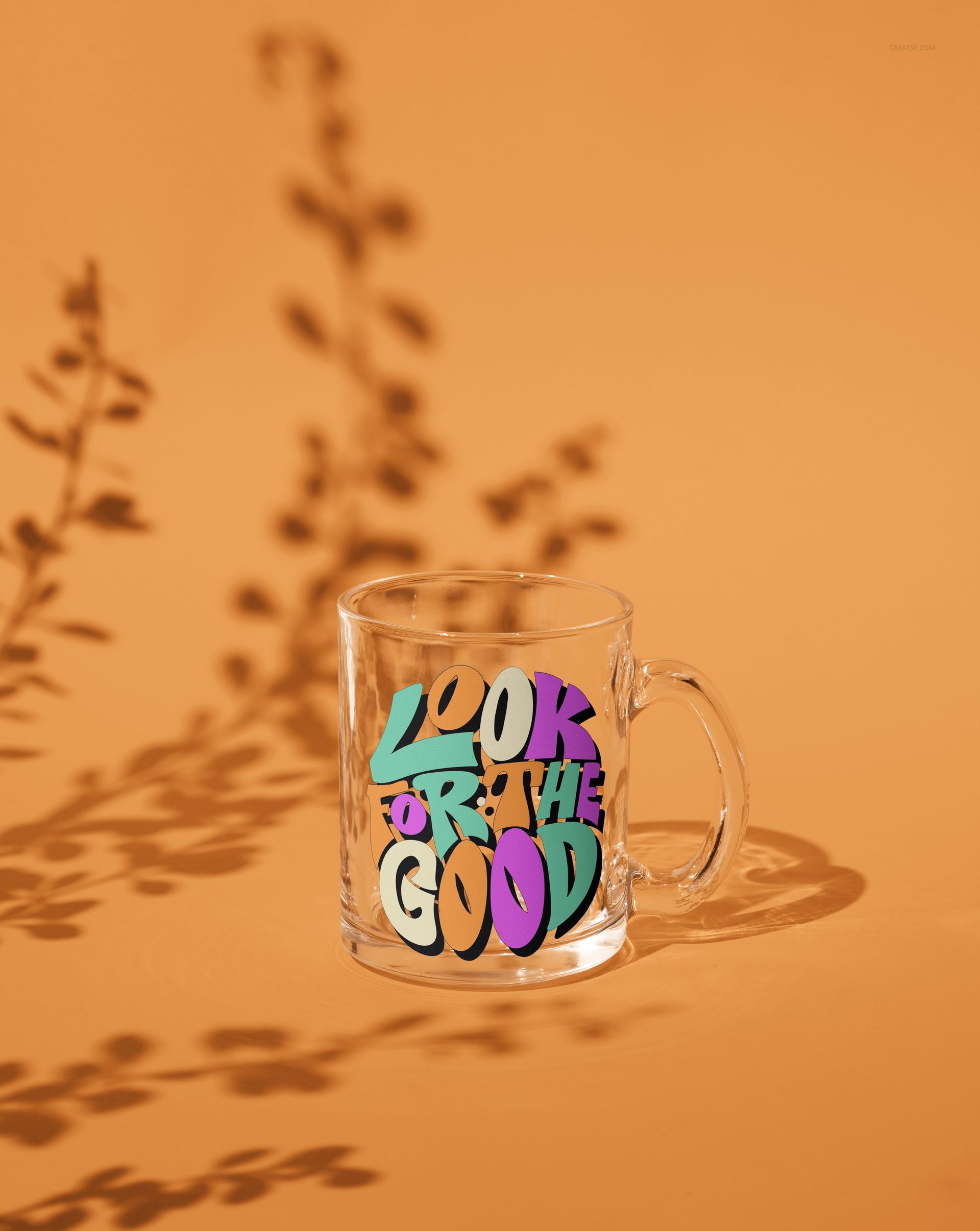 Glass mug mockup displaying bold multicolored text, photographed from the front with leaf-shaped shadows on orange.