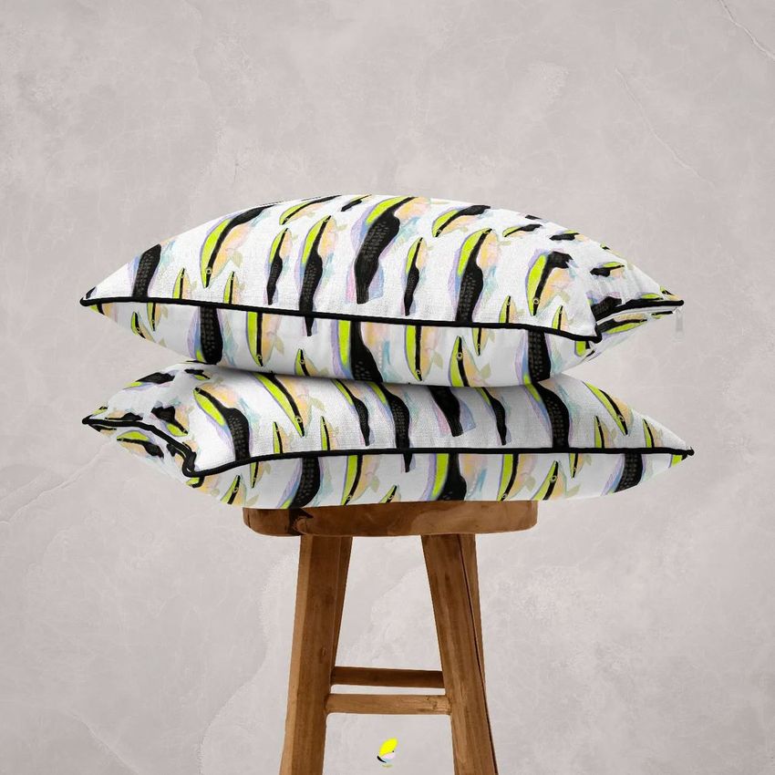 Two reversible piped cushions with a bold abstract feather pattern in black, yellow, and green stacked on a wooden stool.