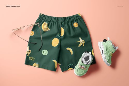 Kids shorts mockup with green fabric featuring banana and pineapple pattern, shown from above on a peach background.