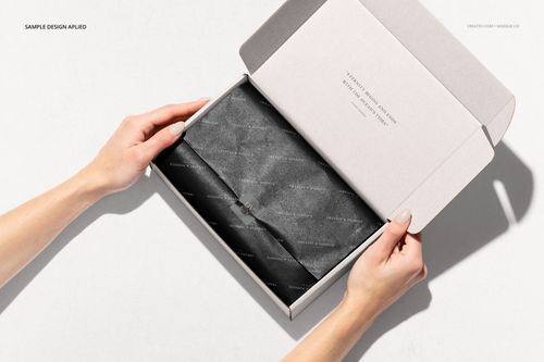 Open packaging box mockup with black tissue paper inside, displayed on a light background with hands holding the box.