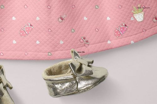 Front view of a baby dress mockup with a quilted pink fabric featuring heart and slipper patterns, next to gold baby shoes.