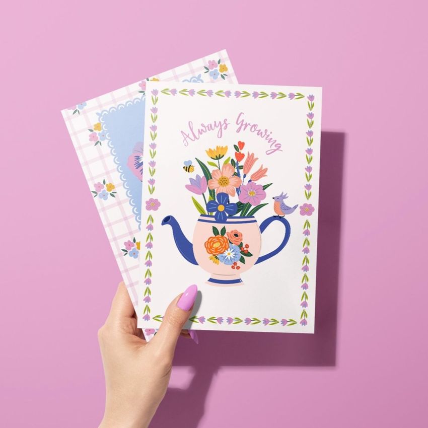 Greeting card mockup showing a hand with pink nails holding two floral cards against a pastel pink background.