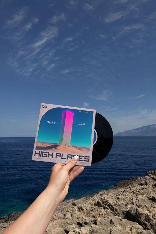 Person’s hand displaying a black vinyl record mockup with vibrant artwork near the ocean and clear sky.