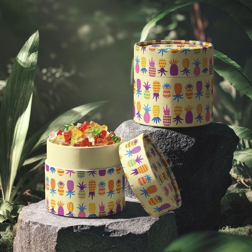 Paper tube packaging mockup with colorful pineapple and jelly designs, shown outdoors on rocks with gummy candies inside.