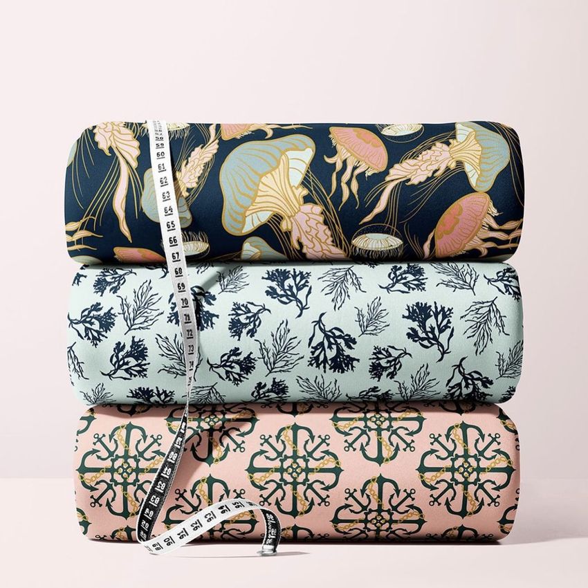 Three rolls of scuba fabric with ocean-inspired patterns are stacked on a light surface, accompanied by a measuring tape.