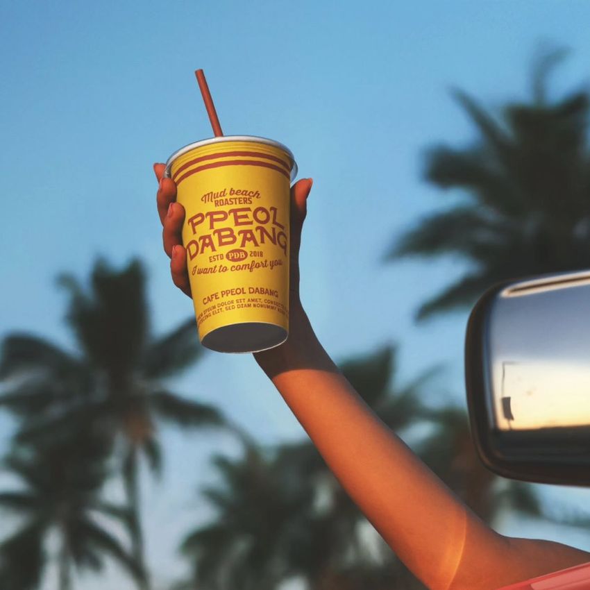 Person’s arm extends from a car, presenting a beverage paper cup mockup against a backdrop of palm trees and sunlight.