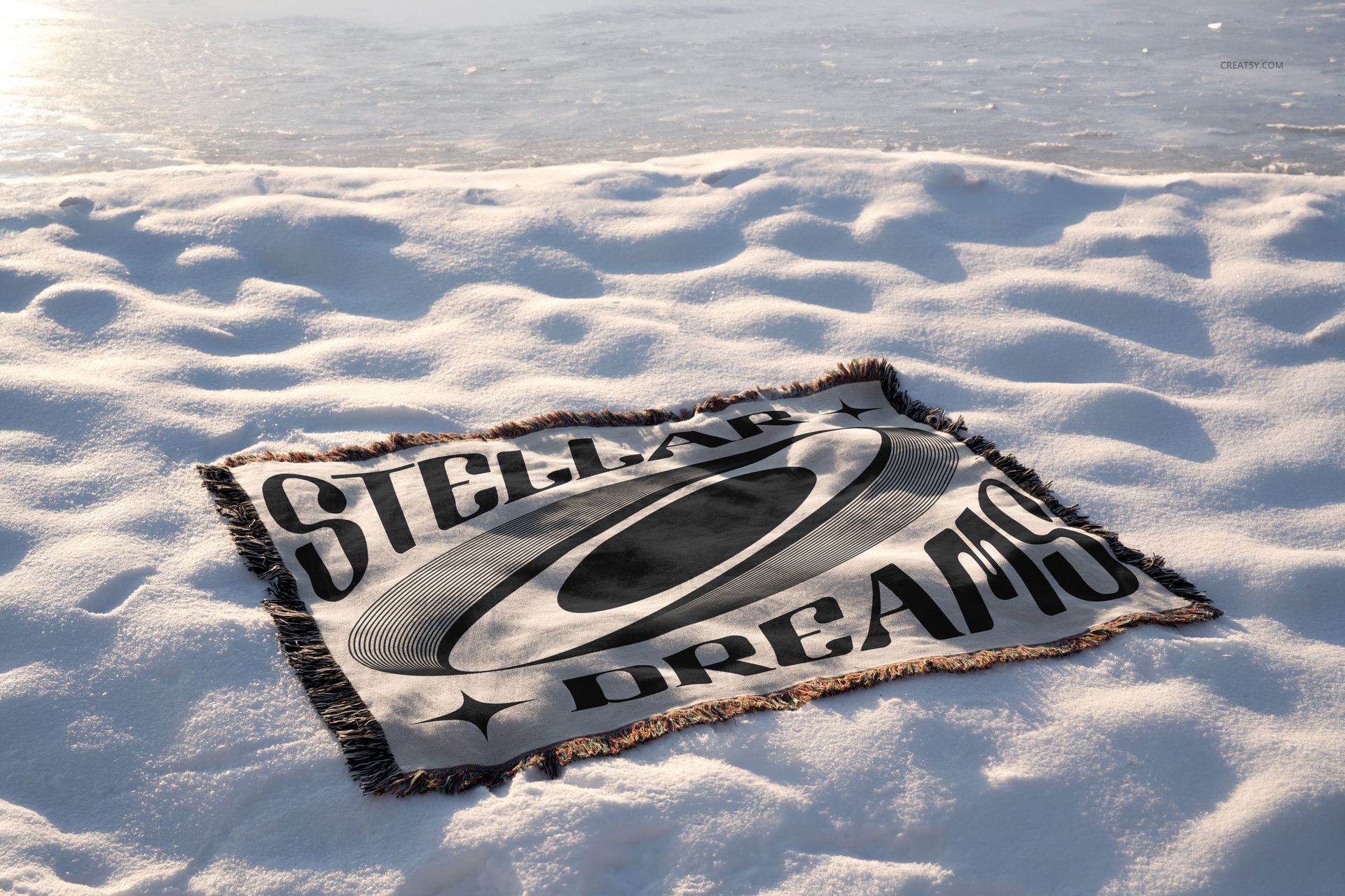 Winter edition blanket mockup showing a fringed textile with graphic print, positioned on a snowy landscape near icy water.