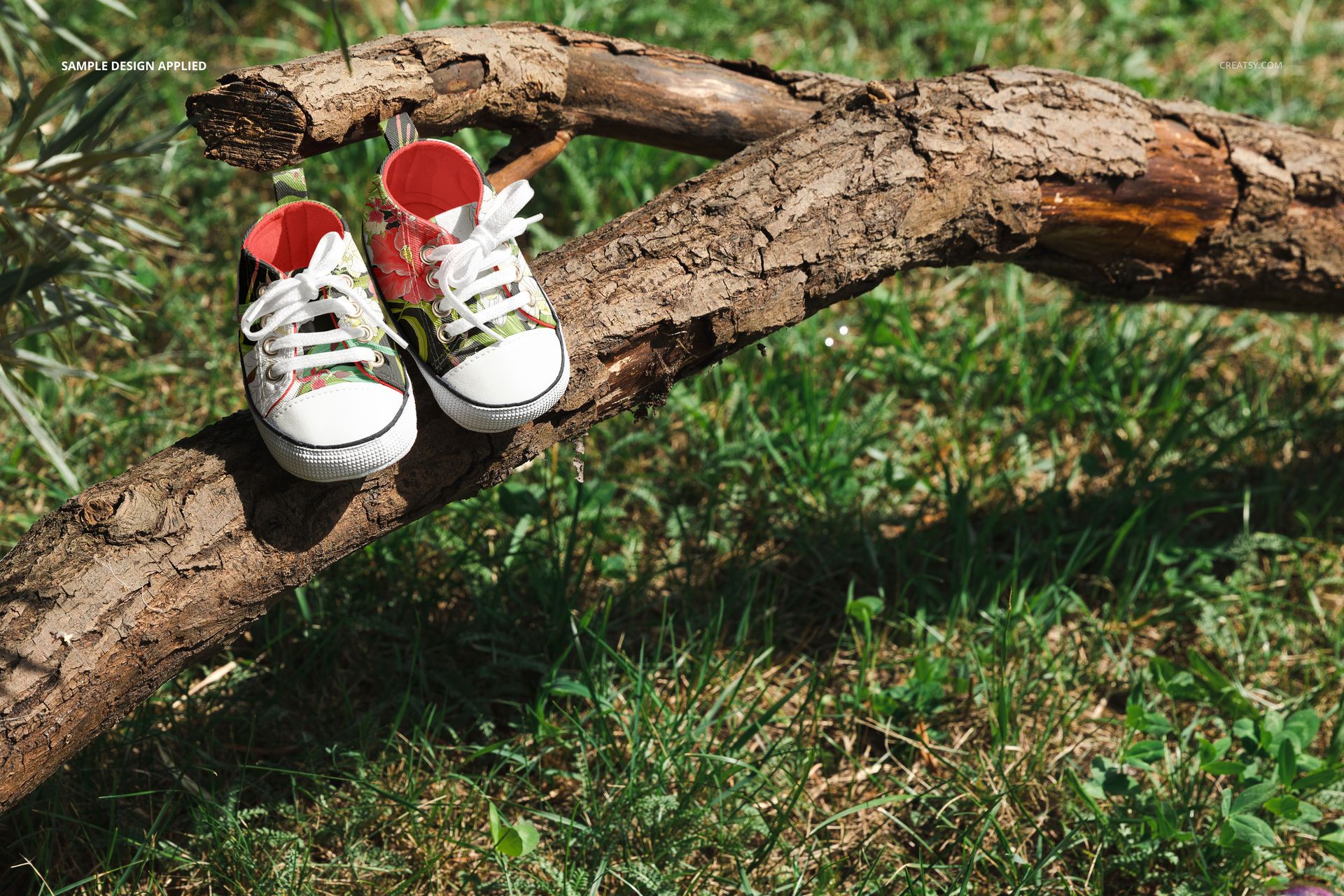 Baby sneakers mockup showing two shoes with vibrant designs placed on a natural tree branch surrounded by green grass.