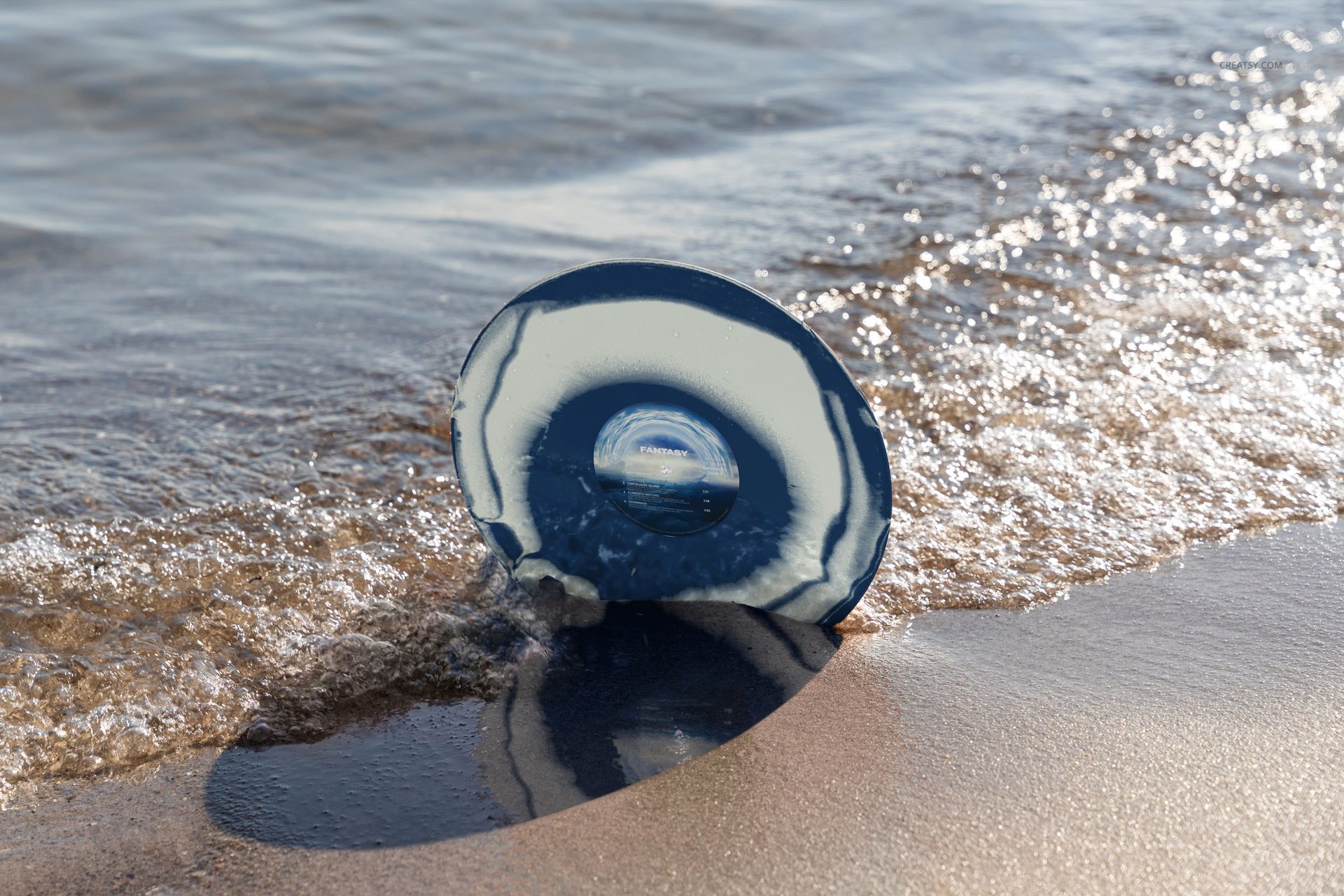 Mockup of a colored vinyl record with a circular blue design resting on sandy shoreline with water washing over it.