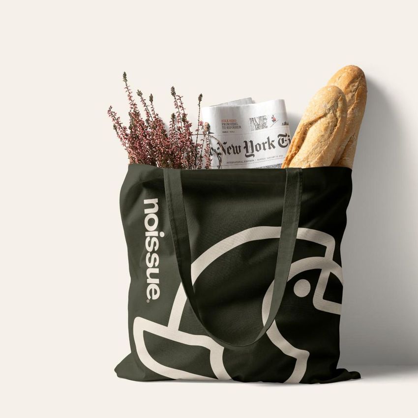 Tote bag mockup featuring a black bag with a bold white graphic, filled with a baguette, newspaper, and sprigs of heather on a light background.