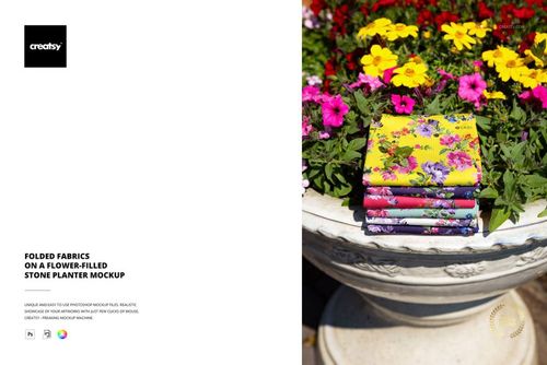 PSD mockup showing multiple folded fabric pieces with floral designs arranged on a flower-filled stone planter.