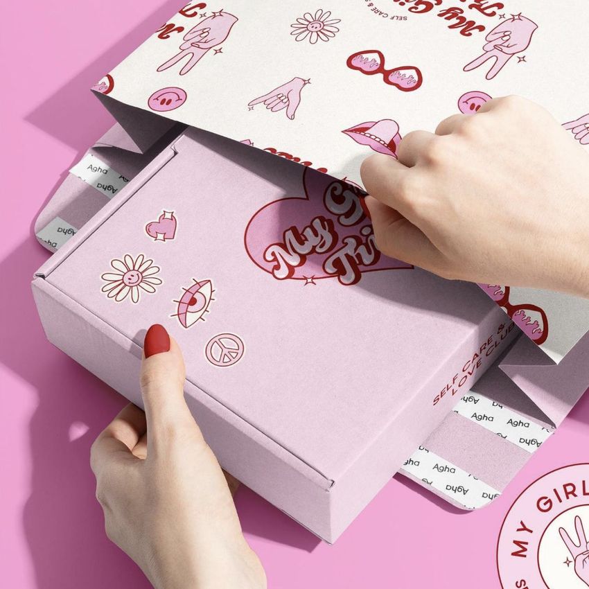 Close-up of hands unboxing a pink mailer mockup featuring fun graphics and text, shown on a solid pink surface.