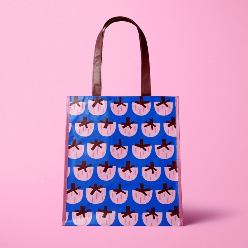 Laminated non-woven tote bag mockup featuring a blue bag with a repeating pink strawberry pattern, set against a pink background.
