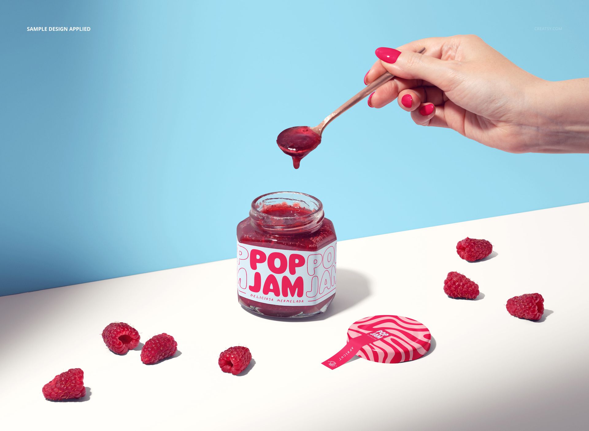 Raspberry jam jar mockup on a white surface with loose raspberries, spoon, and bright blue background.