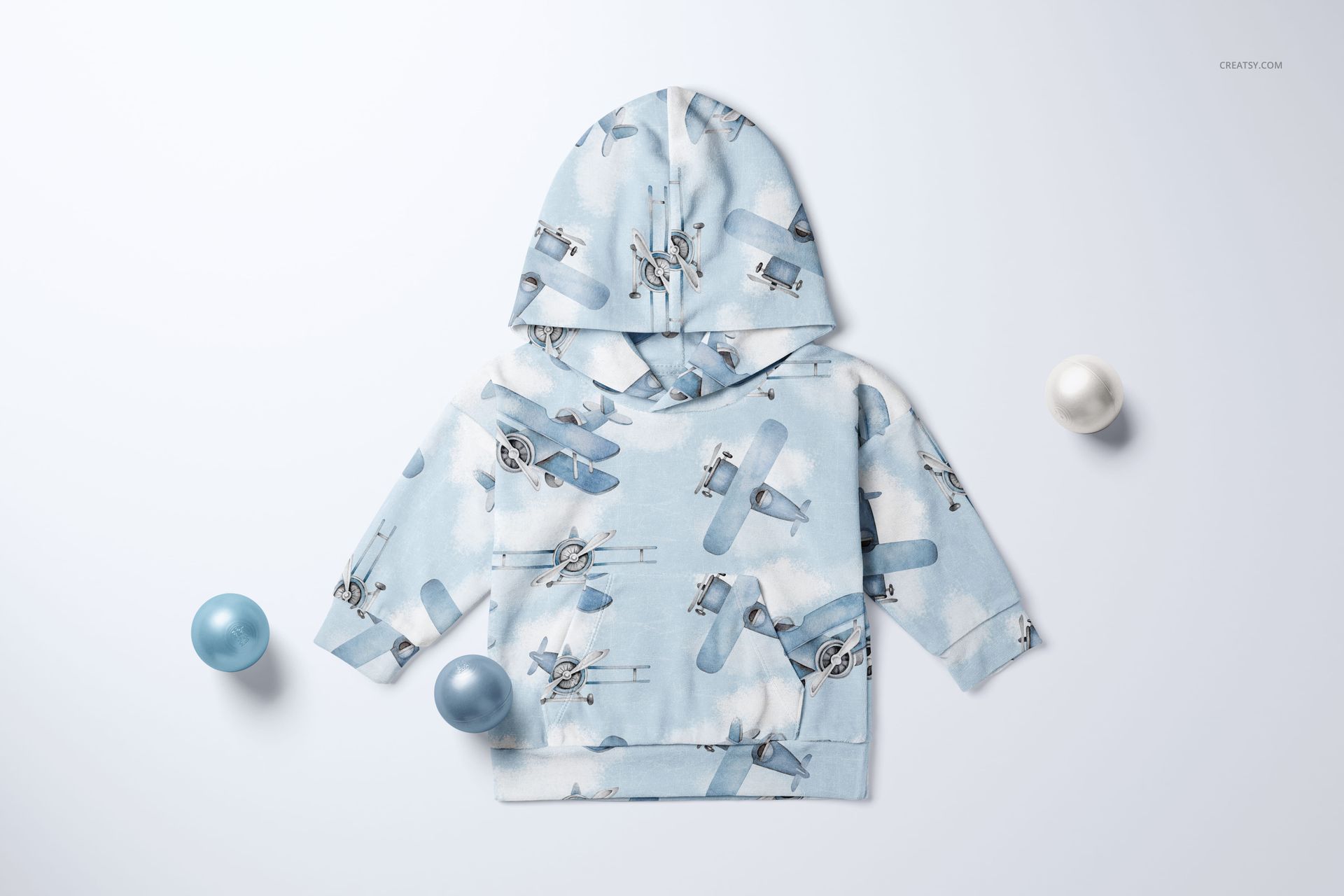 Mockup of a baby velour tracksuit top in pastel blue with aviation print, laid out on a white surface with decorative balls.