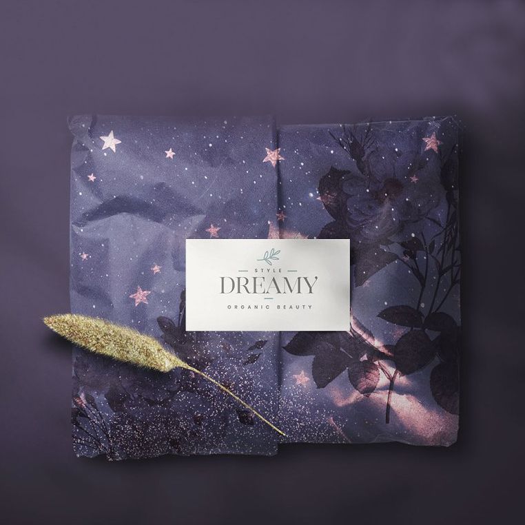 Tissue paper mockup featuring dark blue wrapping paper with a dreamy star and floral pattern, folded neatly with a white label in the center and a dried plant stem on top.