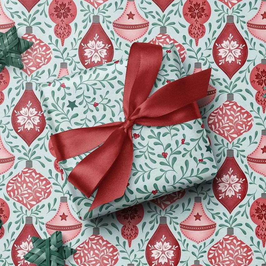 Square gift box mockup wrapped in festive paper with red and green holiday ornament pattern, tied with a wide red ribbon bow.