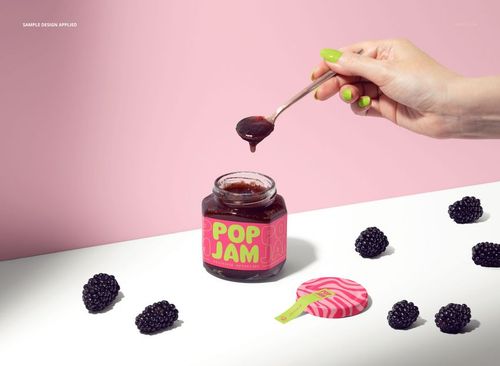 Blackberry jam jar mockup with colorful label, loose blackberries on white surface, and hand holding spoon against pink background.