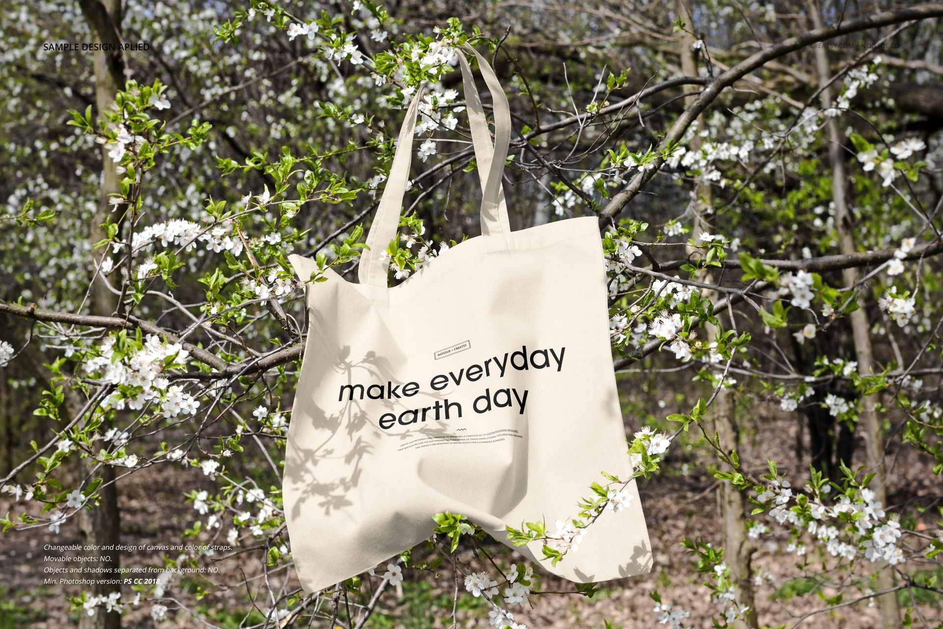Canvas tote bag mockup displayed outdoors, featuring black lettering and surrounded by spring blossoms and branches.