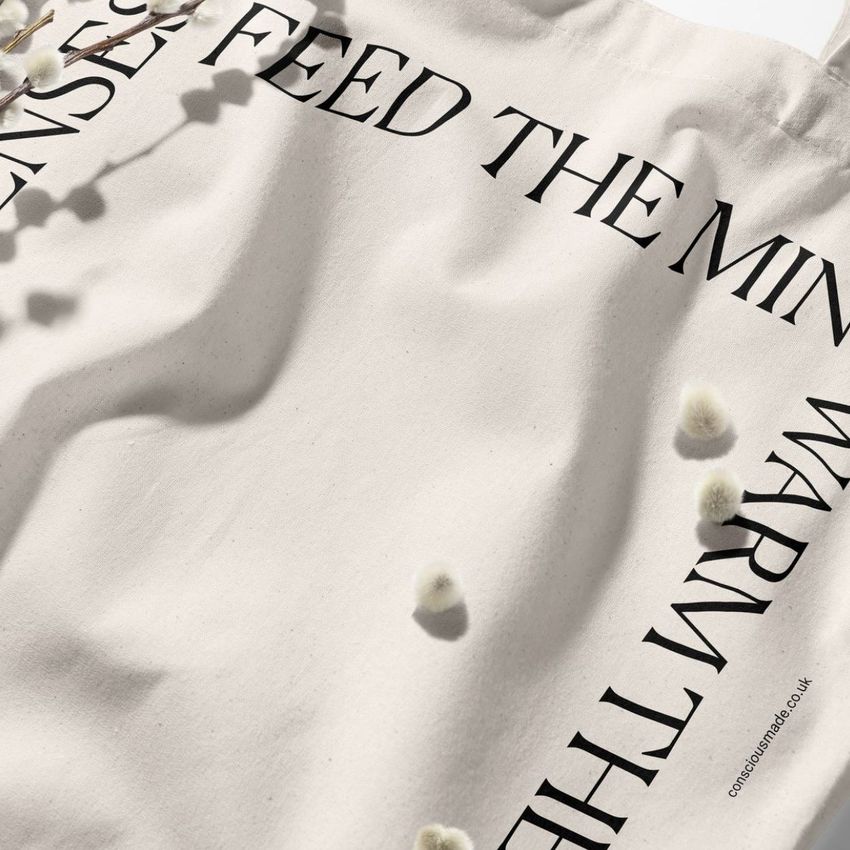 Tote bag mockup featuring black text printed along the edges of a cream fabric, with soft shadows and small white buds.