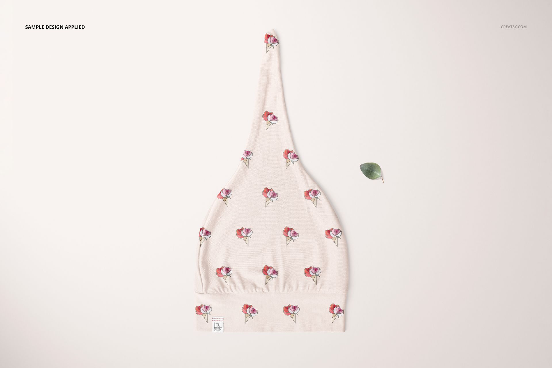 PSD mockup of a baby swaddle hat in a top view, decorated with small floral designs and placed beside a green leaf.