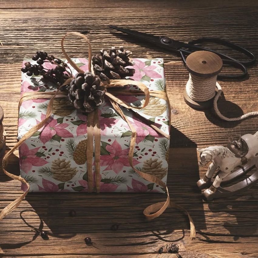 Gift mockup wrapped in floral and pinecone patterned paper, tied with twine and pinecones, on a rustic wooden table.