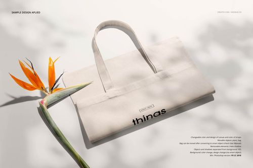 Neutral canvas tote bag mockup lying flat on a light surface, front view with a bird of paradise flower beside it.