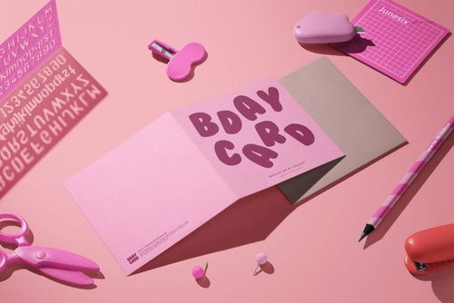 Small square greeting card mockup displayed open on a pink surface, surrounded by stationery items and props.