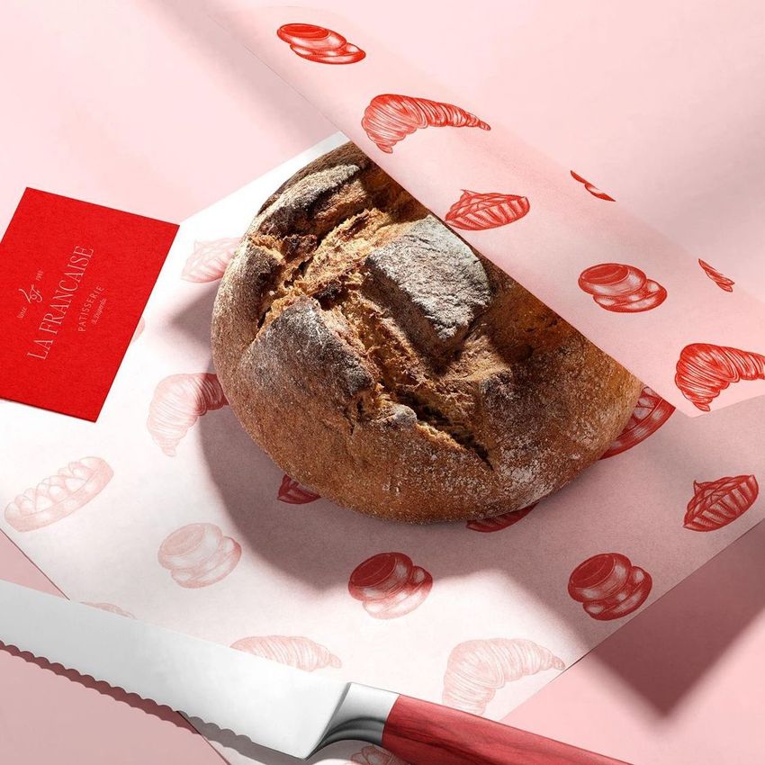 Bakery packaging mockup showing a round loaf of bread wrapped in white food safe paper with red pastry illustrations on a pink surface.