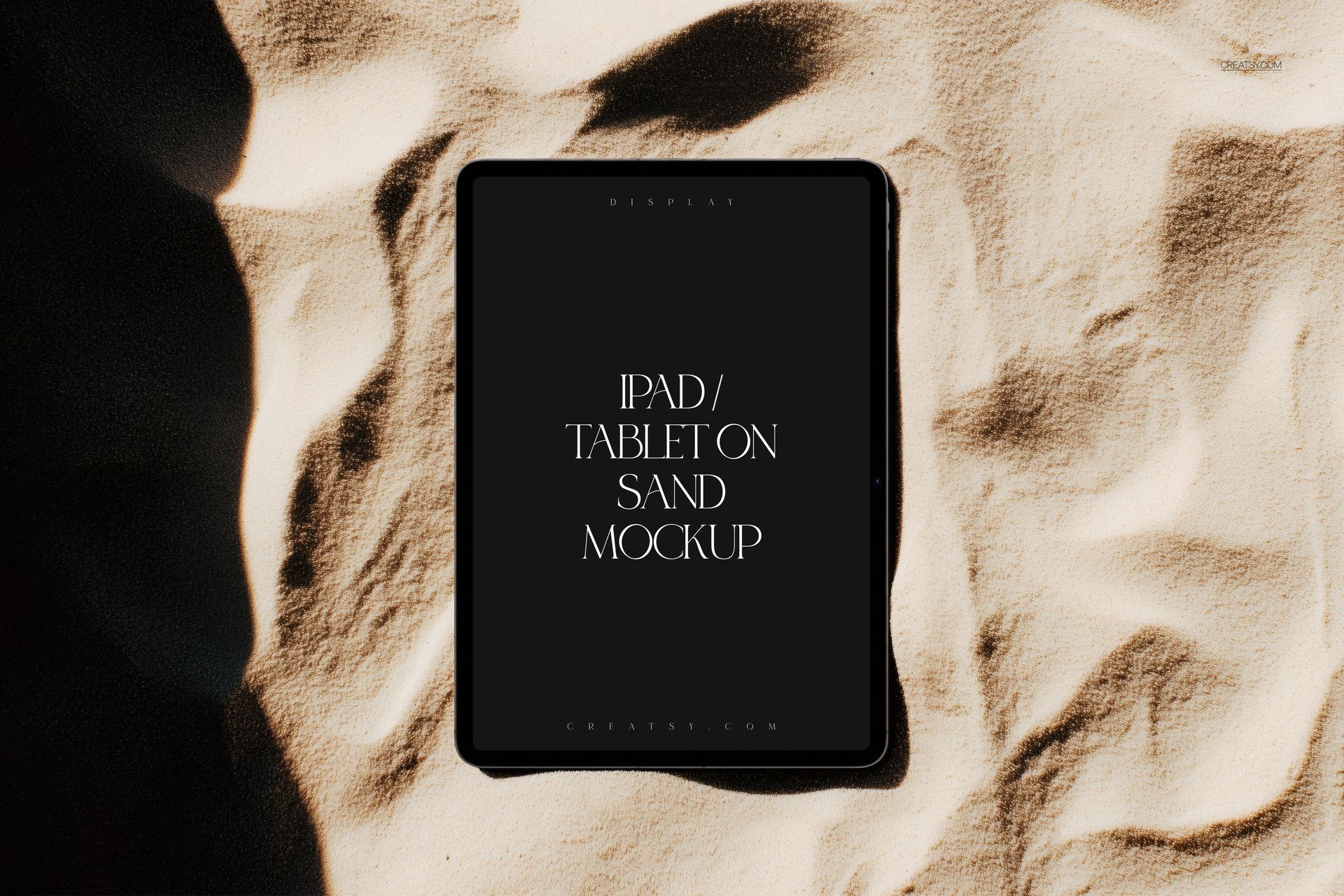 Top view of an iPad Pro mockup resting on light-colored sand, showing a blank dark screen and shadow details.