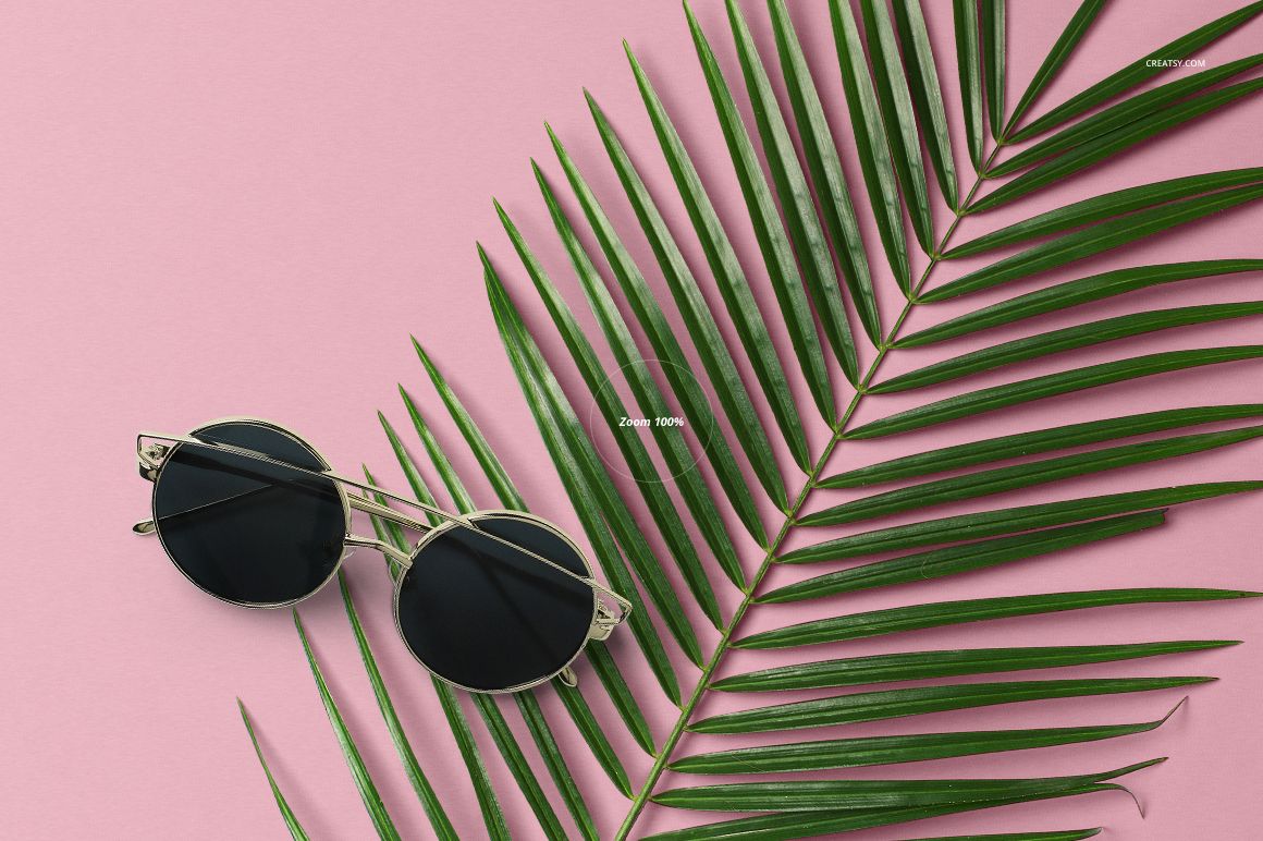 Round sunglasses with dark lenses placed next to a green palm leaf on a pastel pink background, top view.