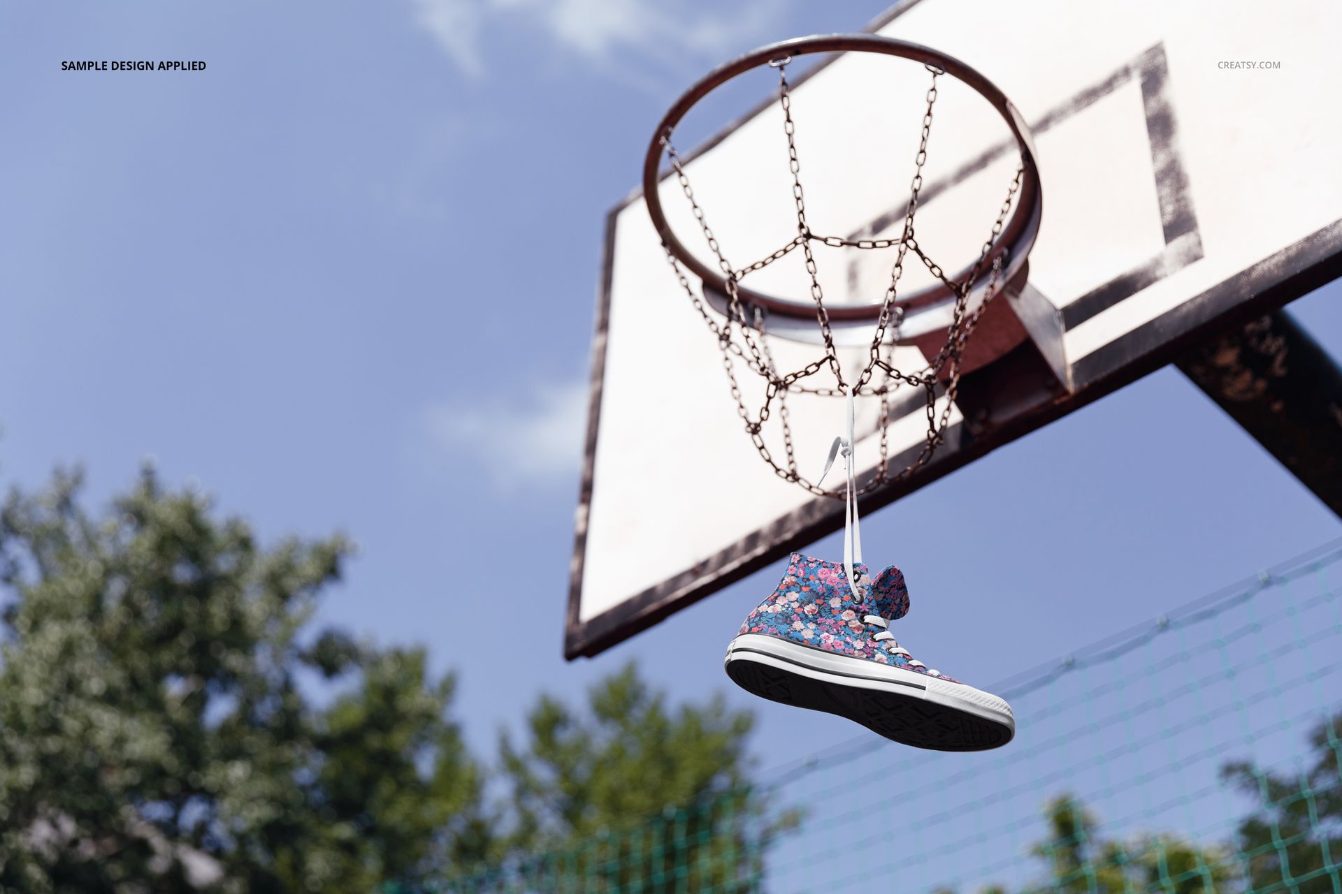 PSD mockup showing a floral print sneaker dangling from a basketball hoop in a sunny outdoor sports setting.