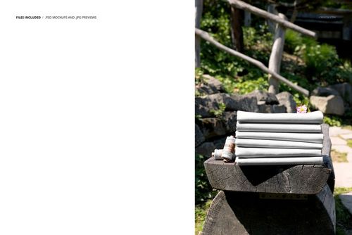 PSD mockup showing a neat stack of plain fabrics placed on a rustic wooden bench outside, with foliage and rocks nearby.