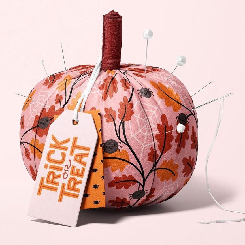 Fabric pumpkin mockup with autumn leaf and spider patterns, used as a pincushion with pins and a tag reading 'Trick or Treat'.