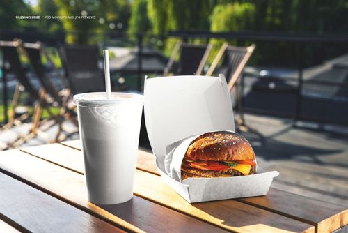 Front view of a burger mockup in a white box next to a plain drink cup on a sunlit wooden table outdoors.