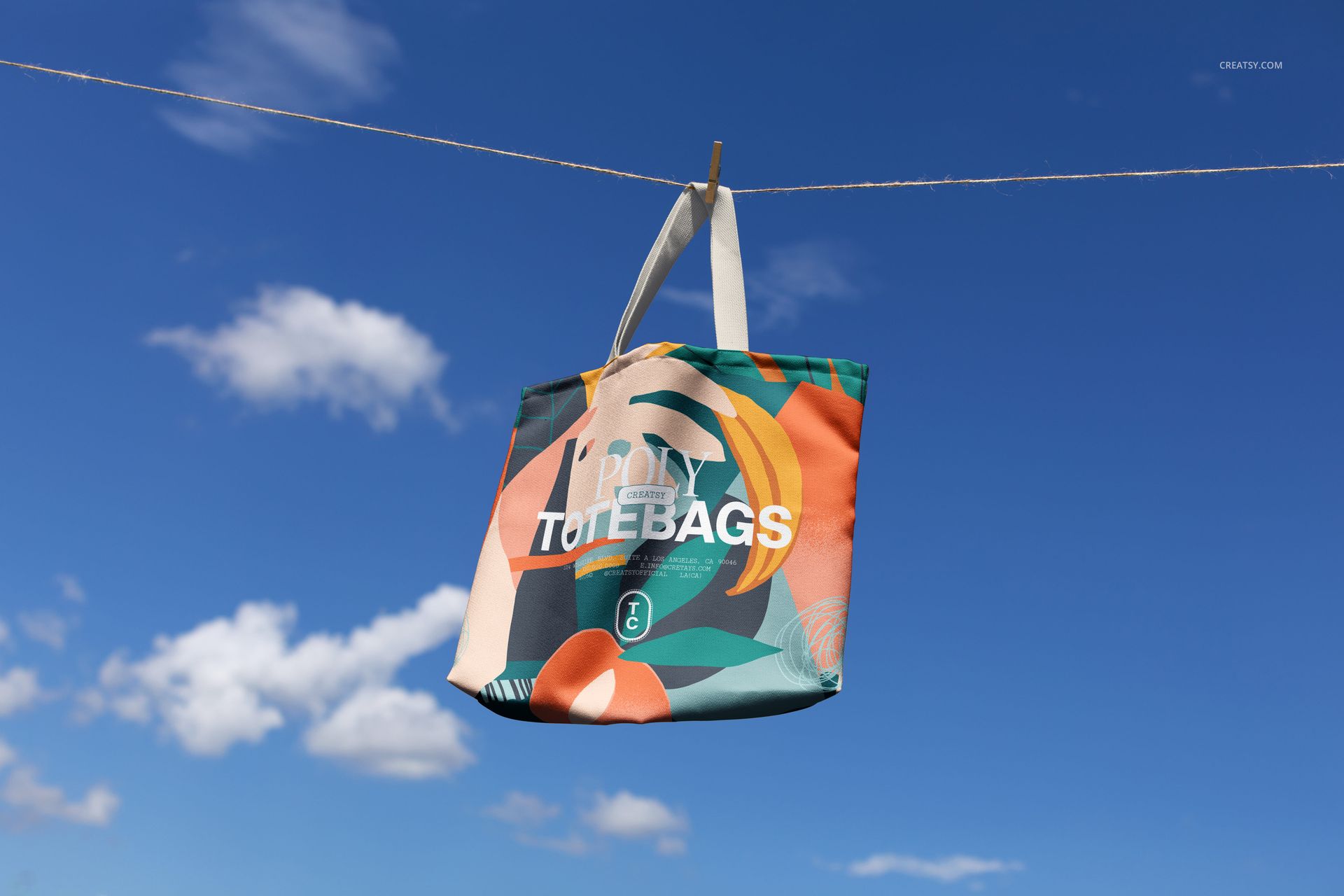 Front view of a large tote bag mockup featuring vibrant geometric designs, suspended outdoors with clouds visible.