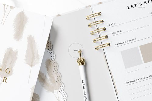 Open spiral-bound planner mockup with beige feather pattern, gold pen, and wedding planning pages in a close-up view.