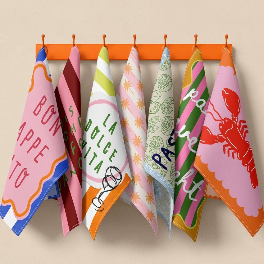 Hanging tea towels mockup showing six colorful towels with various patterns and text, displayed on orange hooks against a beige wall.