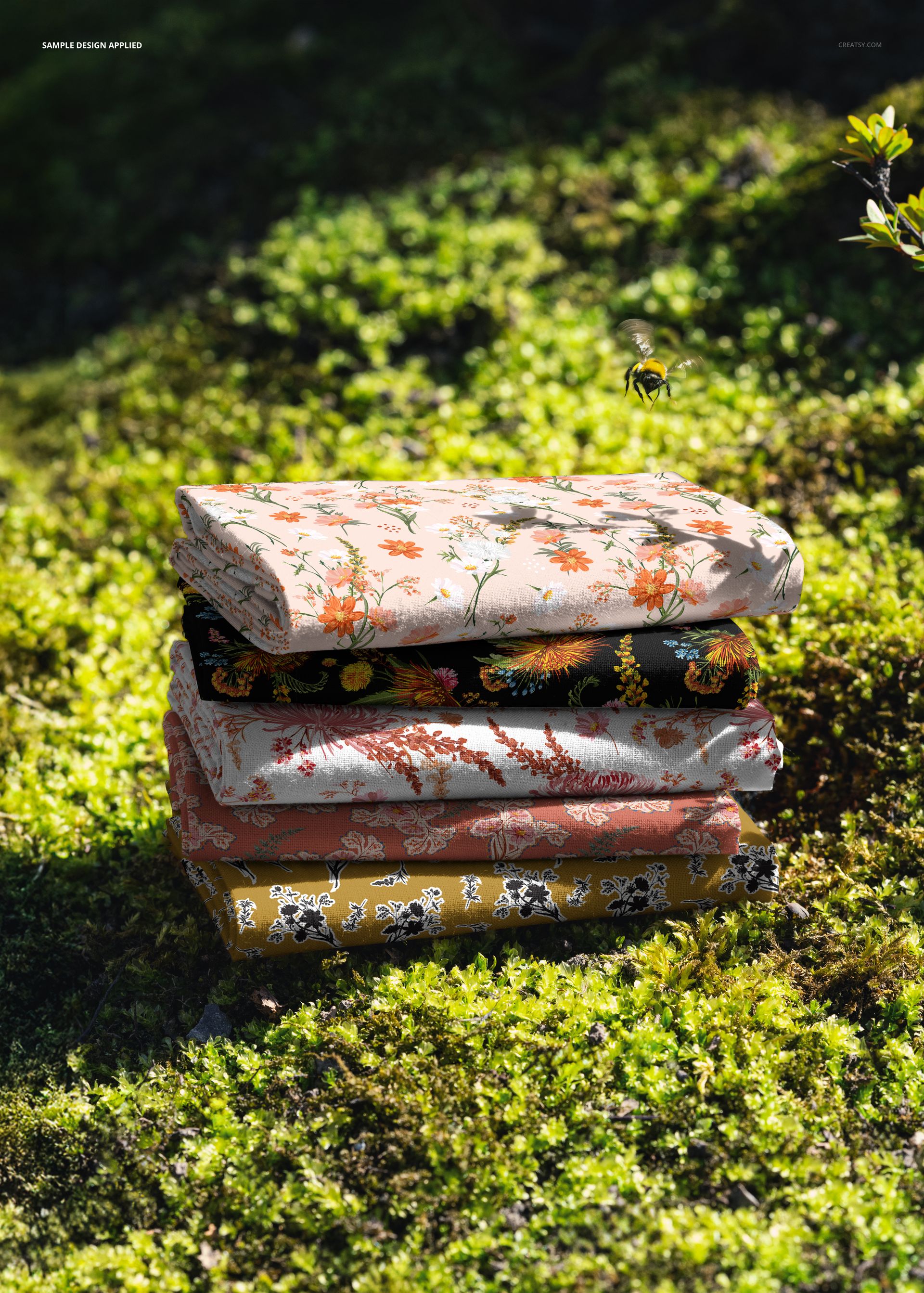PSD mockup showing a pile of colorful floral fabrics resting on vibrant moss with a bee hovering above.