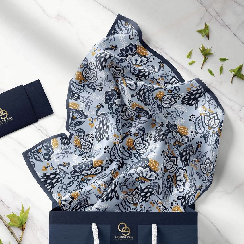 Silk scarf mockup featuring a blue and gold floral peacock pattern draped over the edge of a navy shopping bag on a marble surface.